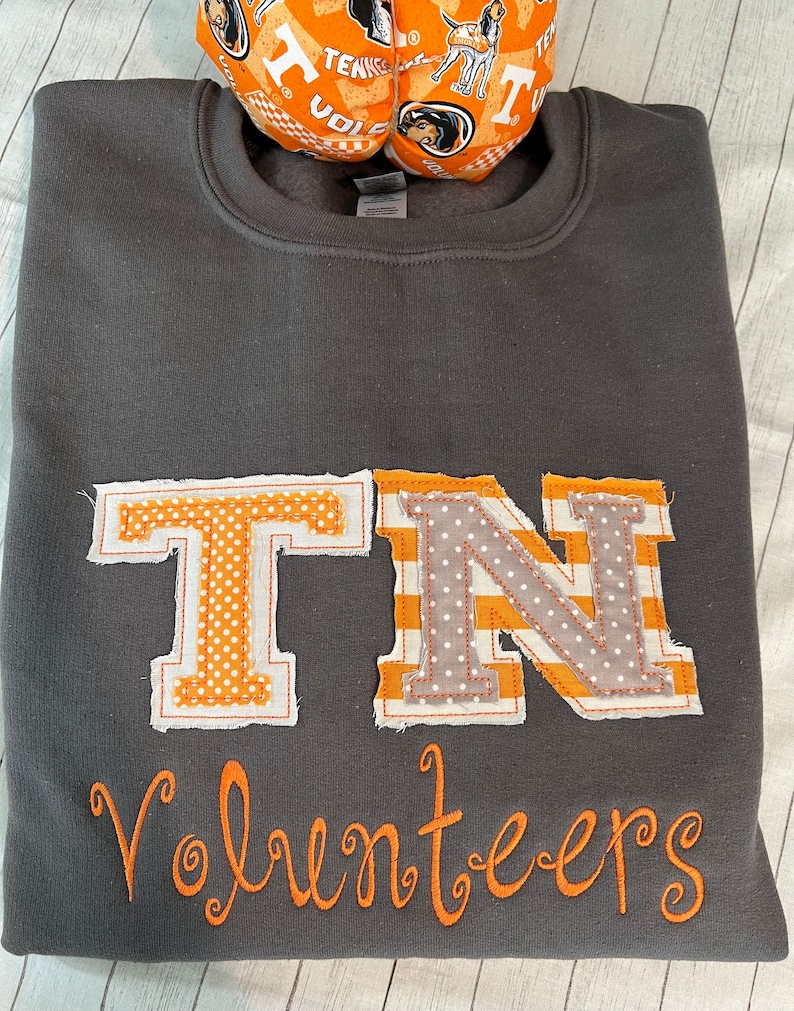 May include: A dark grey sweatshirt with a large orange and white "TN" appliqu&eacute; and the word "Volunteers" embroidered in orange below. The sweatshirt is decorated with a patchwork style design.