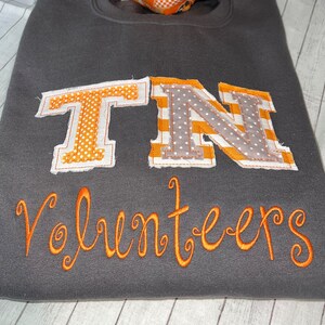 May include: A grey sweatshirt with the letters "TN" in orange and grey fabric, with white polka dots and stripes. The word "Volunteers" is embroidered in orange below the letters.