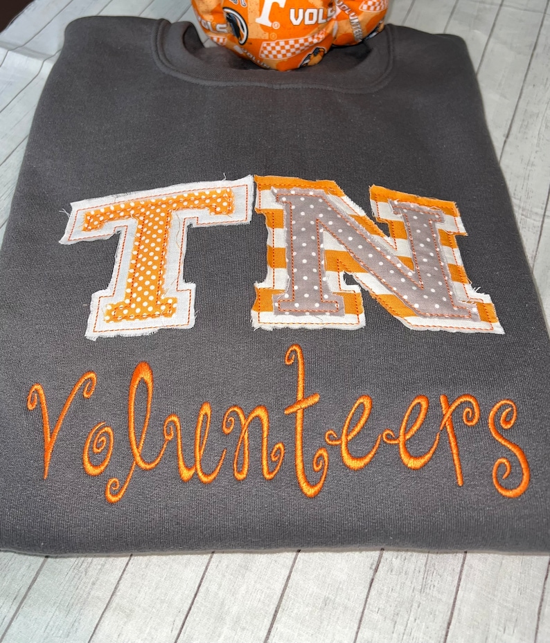 May include: A grey sweatshirt with the letters "TN" in orange and grey fabric, with white polka dots and stripes. The word "Volunteers" is embroidered in orange below the letters.