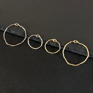 Könnte beinhalten: Vier Schmuckstücke in Form von offenen Kreisen in verschiedenen Größen und metallischen Ausführungen: Silber und Gold. Jeder Ring hat einen strukturierten, organischen Rand und eine kleine Schlaufe oben. Die Teile sind auf einer dunklen Oberfläche angeordnet.