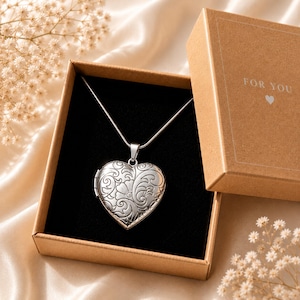 Könnte beinhalten: Eine herzförmige Medaillon-Halskette aus Silber mit einem detaillierten floralen Rankenmuster, platziert in einer braunen Kartonschachtel. Der Deckel trägt den Schriftzug FOR YOU mit einem kleinen Herzsymbol. Das Medaillon hängt an einer Kette.