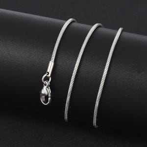 Könnte beinhalten: Eine silberfarbene Schlangenkette mit einem Karabinerverschluss. Die Kette besteht aus kleinen, miteinander verbundenen Gliedern, die eine glatte, flexible Textur erzeugen. Die Halskette wird vor einem dunklen Hintergrund gezeigt.