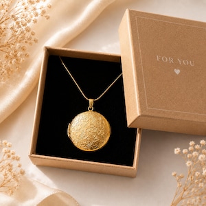 Könnte beinhalten: Eine goldfarbene, runde Medaillon-Halskette mit einer detaillierten Blumen-Gravur auf der Vorderseite, die in einer quadratischen braunen Geschenkbox aus Karton liegt. Der Deckel trägt den weißen Aufdruck FOR YOU mit einem kleinen Herz.