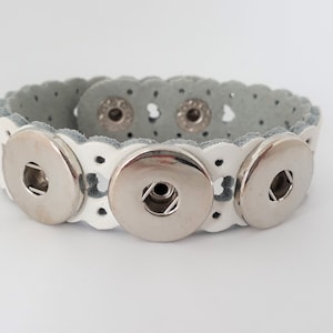 Könnte beinhalten: Weißes Lederarmband mit drei silbernen Druckknöpfen. Das Armband hat einen gewellten Rand und ist verstellbar.