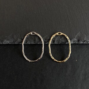 Könnte beinhalten: Zwei ovale Ohrringe, einer silberfarben und einer goldfarben, mit gehammerter Textur. Die Ohrringe werden auf einer dunklen, strukturierten Oberfläche präsentiert. Das Design ist schlicht und elegant.