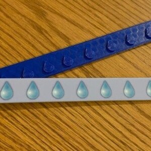 Peut inclure: Deux règles en plastique de couleur bleue et blanche. La règle blanche a une rangée de gouttes de pluie bleues imprimées dessus. La règle bleue a une rangée de bosses en relief.