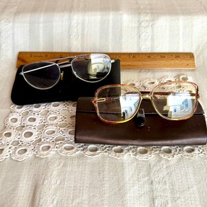 May include: Two pairs of eyeglasses rest on cases. One pair has a silver frame and the other has a gold frame. The glasses are on a white lace doily and a wooden ruler.