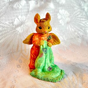 May include: A ceramic figurine of a mouse angel wearing a green and orange patchwork robe. The mouse has a halo and wings and is holding a piece of the robe in its paws.