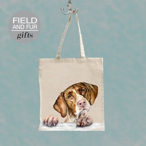 Könnte beinhalten: Eine weiße Canvas-Tragetasche mit braunen Henkeln. Die Tasche zeigt eine Aquarell-Illustration eines braun-weißen Deutsch Kurzhaar-Hundes, der über den oberen Rand der Tasche schaut. Der Text "FIELD AND FUR gifts" ist in Grau auf einem weißen Kreis in der oberen linken Ecke der Tasche gedruckt.