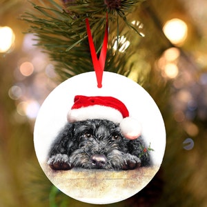 Könnte beinhalten: Runder Weihnachtsschmuck mit einem schwarzen Hund, der einen roten Weihnachtsmannhut mit weißem Rand trägt. Der Hund schaut den Betrachter mit einem lieben Ausdruck an. Ein rotes Band ist zum Aufhängen befestigt. Durchmesser ca. 8 cm.