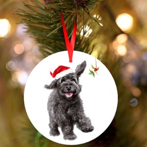 Könnte beinhalten: Runder Weihnachtsschmuck mit einer Aquarellillustration eines schwarzen Hundes mit Weihnachtsmütze. Der Hund lächelt. Ein rotes Band ist zum Aufhängen befestigt. Ein Stechpalmenzweig befindet sich auf der rechten Seite des Ornaments.
