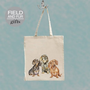 Puede incluir: Una bolsa de lona blanca con el texto "FIELD AND FUR gifts" impreso en la parte superior. La bolsa presenta una ilustración de acuarela de tres perros dachshund.