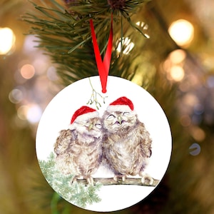 Peut inclure: Ornement de Noël rond avec deux hiboux portant des bonnets de Père Noël, perchés sur une branche avec du feuillage vert. Les hiboux ont les yeux fermés. Un ruban rouge est attaché pour l'accrocher. Diamètre d'environ 8 cm.