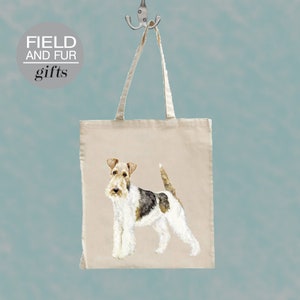 Op de afbeelding: Een witte canvas draagtas met een bruine, witte en zwarte illustratie van een Wire Fox Terrier hond. De tas heeft zwarte handvatten en de tekst "FIELD AND FUR gifts" is in zwart gedrukt op een witte cirkel in de linkerbovenhoek.