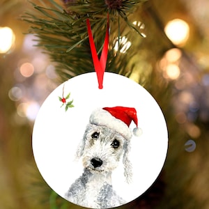 May include: Circular Christmas decoration with a watercolour illustration of a Bedlington Terrier wearing a Santa hat. The ornament has a red ribbon and a sprig of holly. The dog has grey fur and a sweet expression.