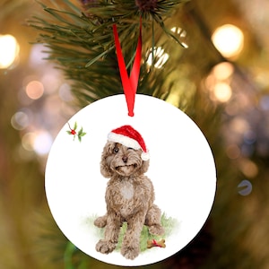 May include: A circular Christmas decoration with a watercolour illustration of a brown dog wearing a Santa hat. The dog is sitting on green grass with a bone. A red ribbon is attached for hanging.