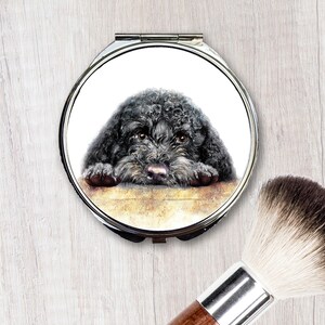 Op de afbeelding: Een compact spiegel met een zwart-witte illustratie van een zwarte poedel die naar de kijker kijkt. De hond rust zijn hoofd op een oppervlak.