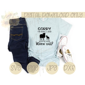 May include: A light blue t-shirt with a black graphic design that says "Sorry for what I said when we were workin' sheep". The graphic includes two black sheep silhouettes. The t-shirt is laid out on a white surface with a pair of blue jeans and white sneakers.