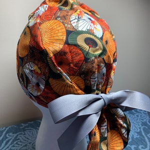 May include: A patterned scrub cap featuring a repeating design of orange, yellow, and white parasols on a dark background. The cap is tied with a gray ribbon bow. The cap is displayed on a white mannequin head.