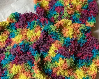 May include: Three colorful crocheted scrubbers in shades of yellow, orange, red, purple, blue, and green. The scrubbers are made of a fluffy, textured yarn.