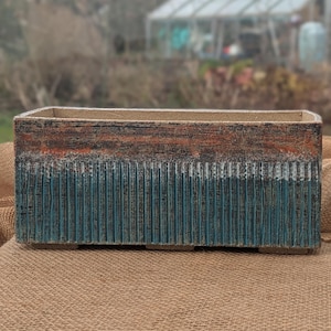 May include: Rectangular planter with a textured surface. The top section is brown and orange, transitioning to a teal blue lower section with vertical grooves. The planter is designed for outdoor use.