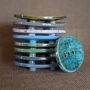 Op de afbeelding: Een stapel keramische zeepschalen in verschillende tinten blauw, groen en wit met gespikkelde patronen. De bovenste schaal is effen teal van kleur met drie gaten.