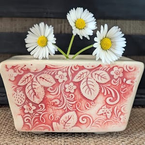 May include: A white ceramic planter with a red floral design and three white daisies in it.