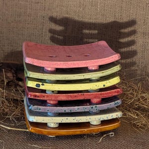May include: A stack of seven rectangular ceramic soap dishes with curved edges and drainage holes. The dishes feature various speckled and solid glaze finishes in colours including pink, yellow, red, purple, blue, cream, and brown, each with small feet.