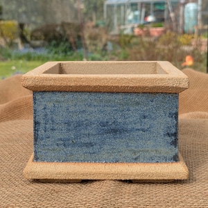 May include: A square ceramic planter with a textured, light brown rim and base. The sides are a mottled blue with a textured finish. The planter is on a burlap surface, with a blurred outdoor background.