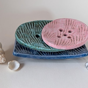 Pode incluir: Um conjunto de três saboneteiras de cerâmica em azul, verde e rosa. Os pratos são empilhados uns sobre os outros, com o prato rosa no topo. Os pratos têm uma superfície texturizada e são projetados para conter sabonete.