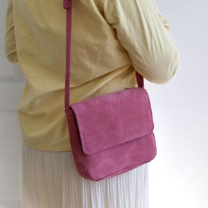 Può includere: Una borsa a tracolla in camoscio rosa con chiusura a patta e tracolla regolabile. La borsa è di una vibrante tonalità di rosa e sembra essere realizzata in morbido materiale scamosciato. La borsa è indossata a spalla.