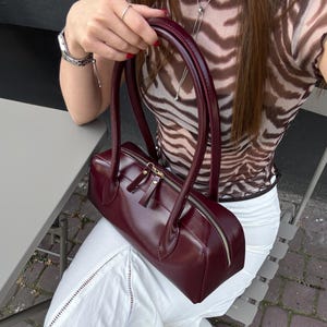 Puede incluir: Un bolso rectangular burdeos con cierre de cremallera y herrajes dorados. El bolso tiene dos asas y lo lleva una persona con un top de estampado de cebra y una falda blanca. El bolso mide unos 30 cm de largo.