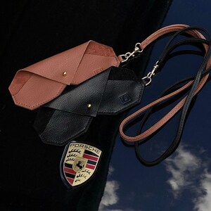 May include: Two leather eyeglass cases, one brown and one black, with a brown and black lanyard. The cases are folded and have a gold metal clasp. The lanyard has a gold metal clip. The cases are on a black surface with a Porsche logo reflected in the surface.