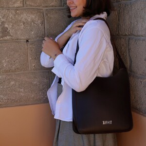 May include: A dark brown shoulder bag with a rectangular shape and a small silver-colored logo. The bag is carried by a person wearing a white shirt and a plaid skirt. The bag has a simple, elegant design.