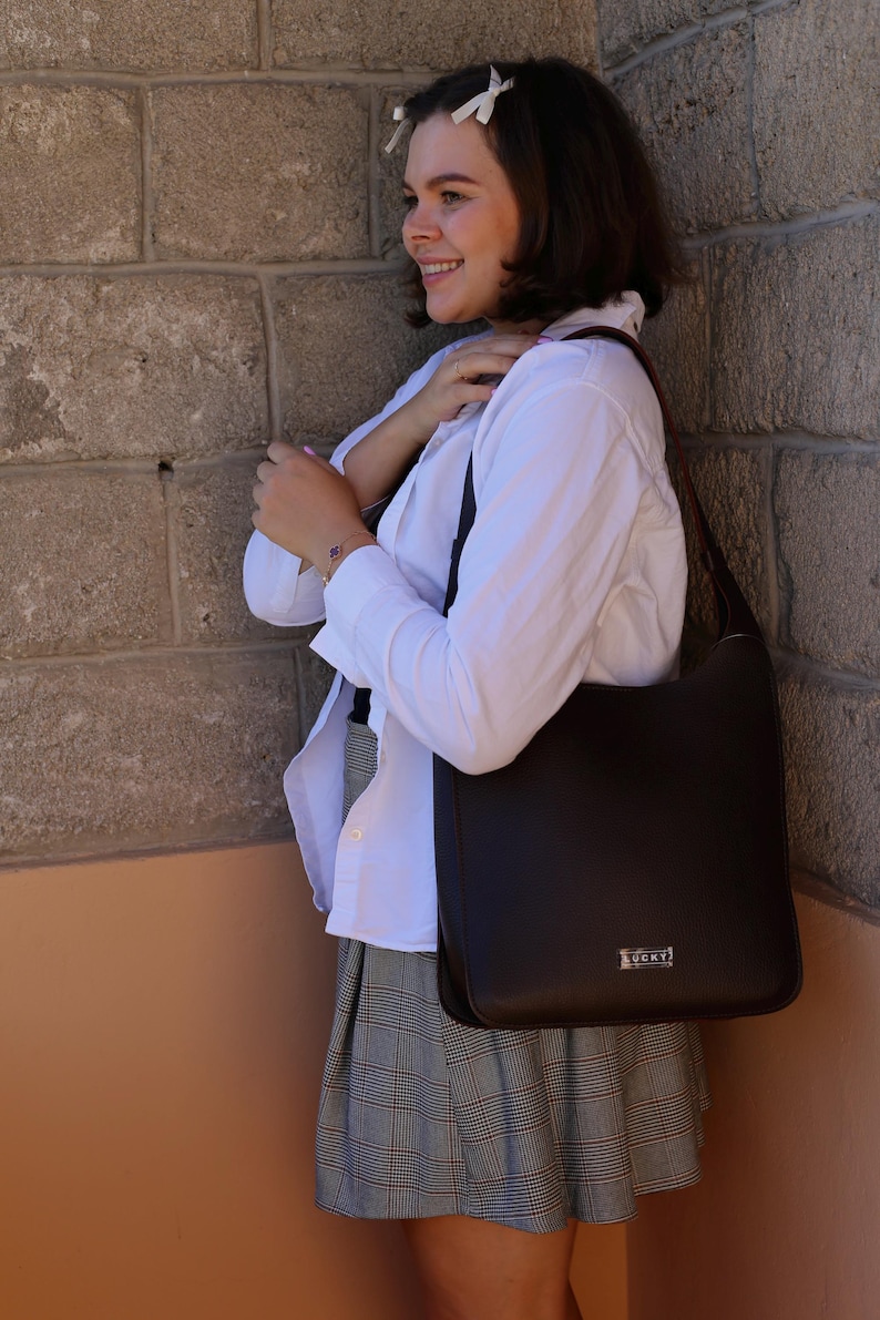 May include: A dark brown shoulder bag with a rectangular shape and a small silver-colored logo. The bag is carried by a person wearing a white shirt and a plaid skirt. The bag has a simple, elegant design.