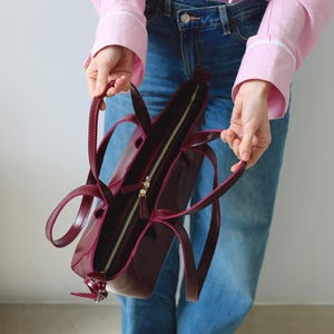 May include: A burgundy leather tote bag with a gold zipper and handles. The bag is open, revealing the interior. The bag is held by a person wearing a pink shirt and blue jeans. The bag is a stylish accessory.