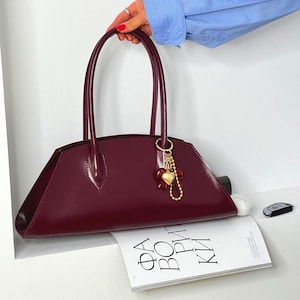 May include: A burgundy handbag with a glossy finish, featuring two arched handles and a gold-toned charm with a heart pendant. The bag is rectangular with a trapezoidal shape and is resting on a magazine.