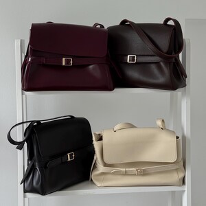 May include: Four structured shoulder bags with fold-over flaps and horizontal buckled straps. The collection includes a smooth burgundy bag, a textured dark brown bag, a smooth black bag, and a textured cream bag, each featuring gold-tone hardware.