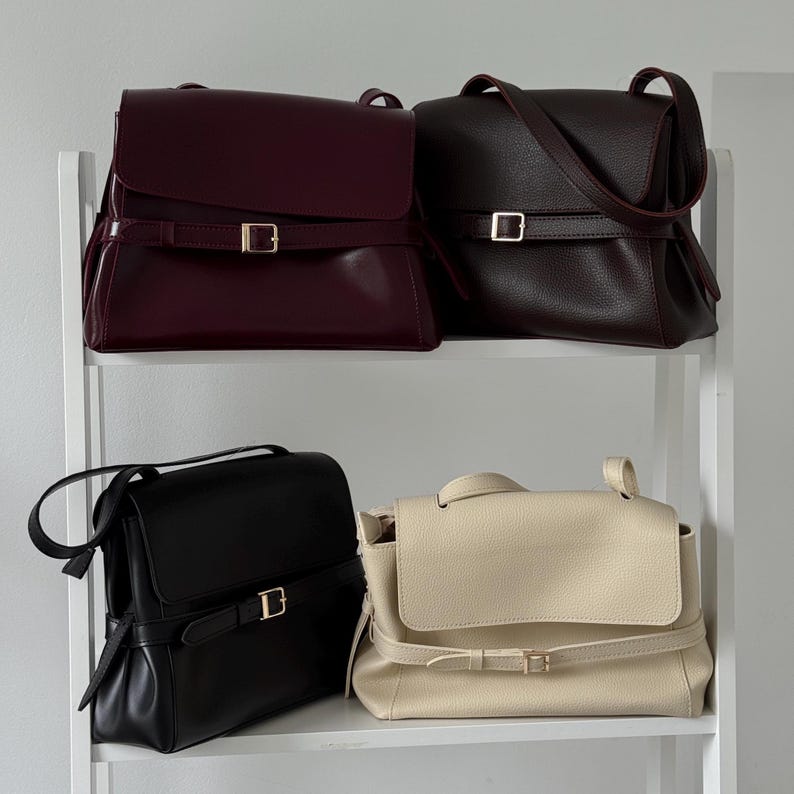 May include: Four structured shoulder bags with fold-over flaps and horizontal buckled straps. The collection includes a smooth burgundy bag, a textured dark brown bag, a smooth black bag, and a textured cream bag, each featuring gold-tone hardware.