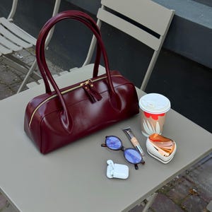 May include: A deep burgundy barrel-shaped handbag with gold-tone zippers and dual handles. Beside it are blue-tinted sunglasses, a small tube of concealer, a white wireless earbud case, a hairbrush, and a paper coffee cup with a white lid.