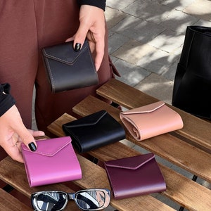 May include: Five different colored leather wallets on a wooden surface. The wallets are all closed and have a flap with a stitched design. The wallets are in shades of black, pink, purple, brown, and beige.