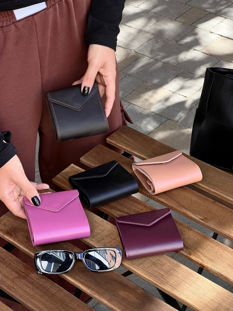 May include: Five different colored leather wallets on a wooden surface. The wallets are all closed and have a flap with a stitched design. The wallets are in shades of black, pink, purple, brown, and beige.
