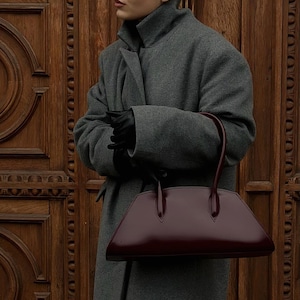 May include: A gray wool coat with a high collar and a burgundy leather handbag. The handbag has a trapezoid shape with a glossy finish and two handles. The person is wearing black gloves and white-framed glasses.