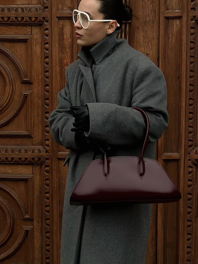May include: A gray wool coat with a high collar and a burgundy leather handbag. The handbag has a trapezoid shape with a glossy finish and two handles. The person is wearing black gloves and white-framed glasses.