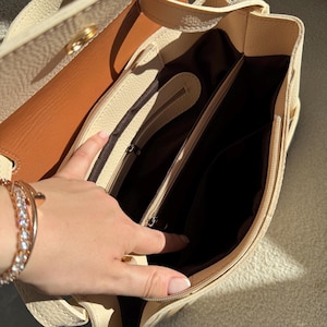 May include: A cream-colored leather handbag with a textured exterior and a smooth tan interior lining. The bag features a central zippered compartment, dark brown fabric lining, and multiple internal pockets for organization with metal zippers.