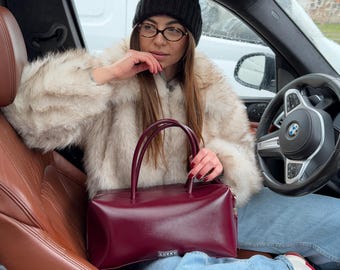 May include: A burgundy leather handbag with a rectangular shape and two top handles. The bag is held in a car, with a person wearing a black beanie, glasses, and a cream-colored faux fur coat. The word "LUCKY" is visible on the bag.