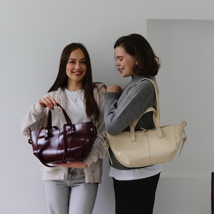 May include: Two women holding leather tote bags. One bag is a deep burgundy color, and the other is a cream color. Both bags have short handles and a structured design. The background is a plain white wall.