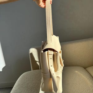 May include: A cream-colored handbag with a textured surface, featuring a single vertical handle and a side strap with metallic studs. The bag has a structured flap closure and a side pocket, showcasing a clean, solid color design throughout.