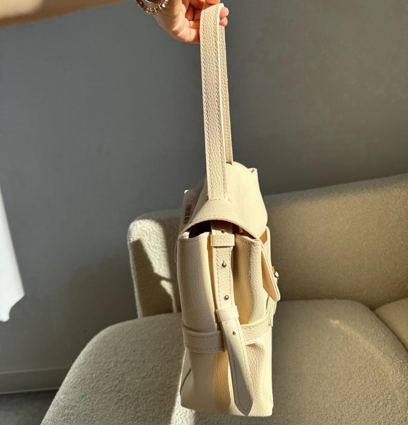 May include: A cream-colored handbag with a textured surface, featuring a single vertical handle and a side strap with metallic studs. The bag has a structured flap closure and a side pocket, showcasing a clean, solid color design throughout.