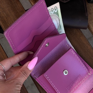 May include: A pink leather wallet with a snap closure and the word "LUSTY" embossed on the front. The wallet is open and shows multiple compartments for cards and cash.